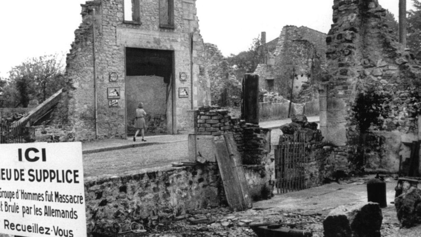 Blick auf die Ruinen von Oradour-sur-Glane und eine Gedenktafel, deren Text lautet: "Gedenkstätte: Eine Gruppe von Männern wurde hier von den Deutschen massakriert und verbrannt. Besinnt Euch"