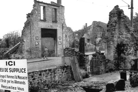 Blick auf die Ruinen von Oradour-sur-Glane und eine Gedenktafel, deren Text lautet: "Gedenkstätte: Eine Gruppe von Männern wurde hier von den Deutschen massakriert und verbrannt. Besinnt Euch"