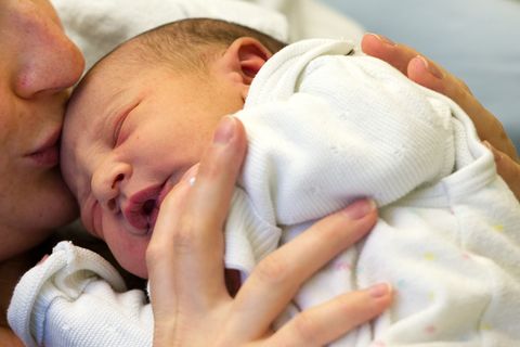 Ein Baby liegt mit geschlossenen Augen auf der Brust der Mutter. Sie hält es fest und küsst seinen Kopf.