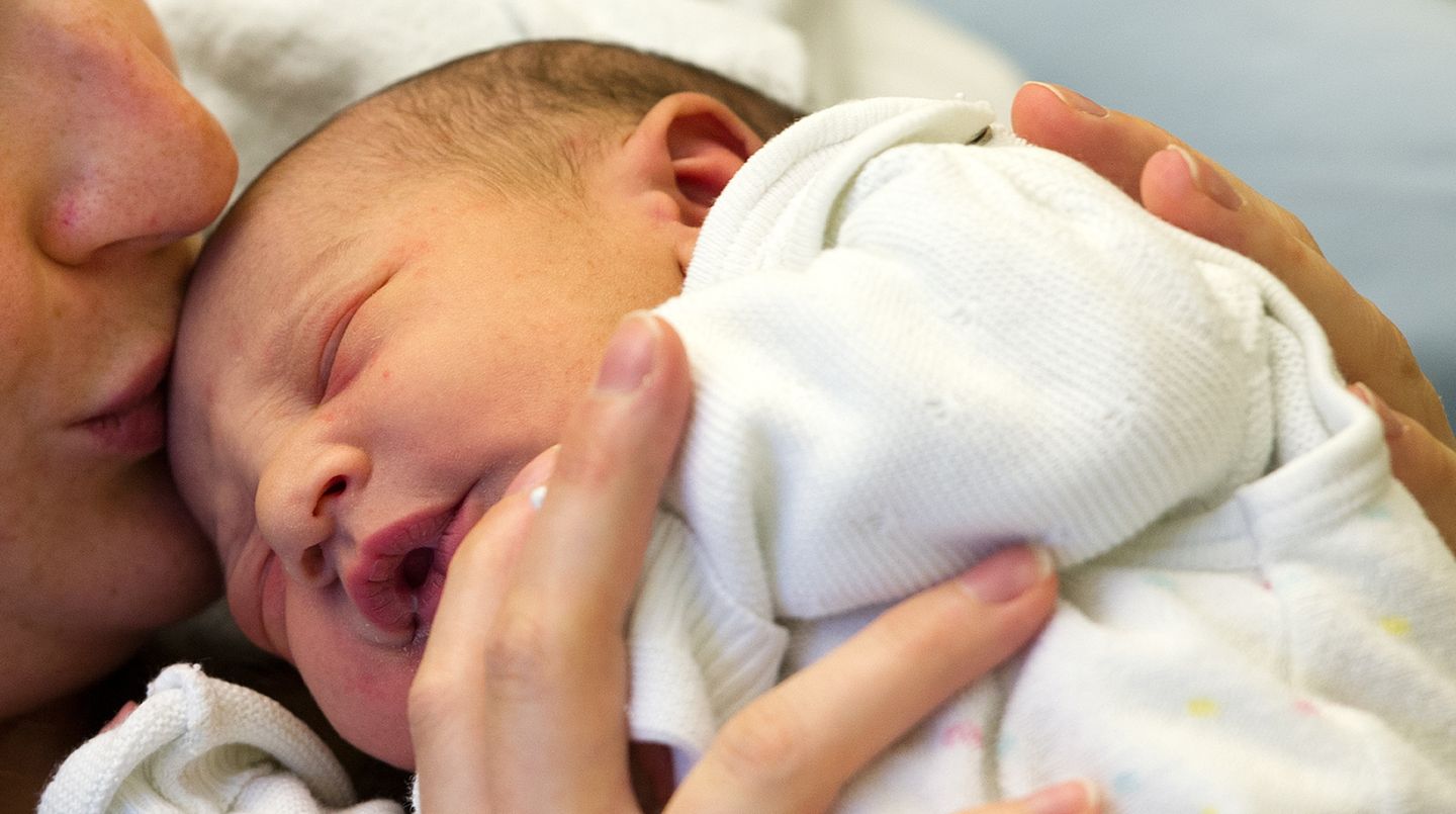 Ein Baby liegt mit geschlossenen Augen auf der Brust der Mutter. Sie hält es fest und küsst seinen Kopf.