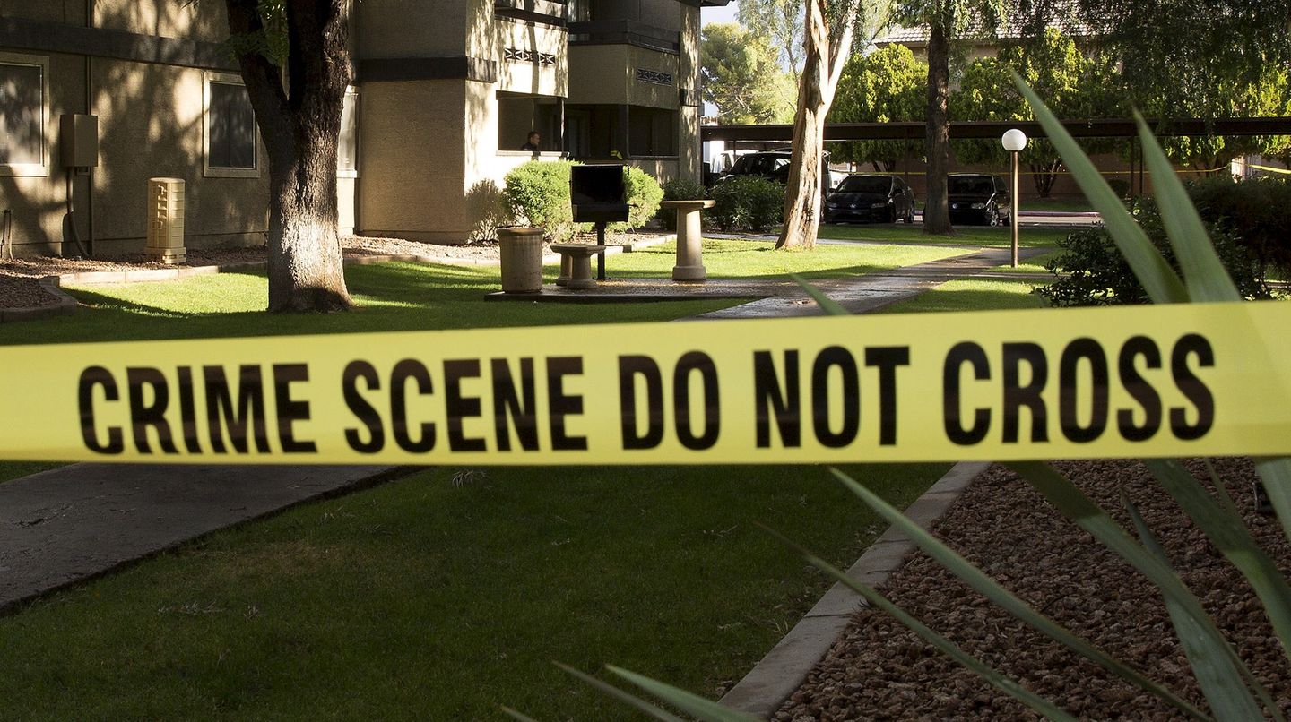 Ein gelbes Polizei-Absperrband mit dem Schriftzug "Crime Scene Do not cross" vor einer Gartenanlage bei Sonnenschein