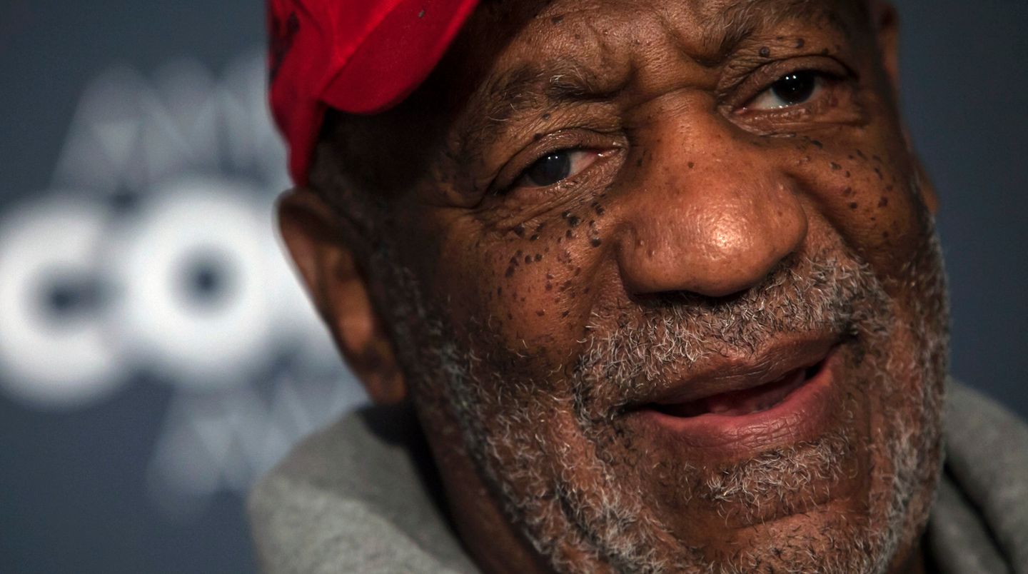 Bill Cosby trägt einen Stoppelbart, eine Schirmmütze und einen Kapuzenpullover. Sein Lächeln wirkt angestrengt.