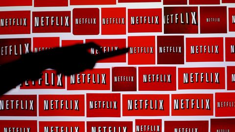 Eine Hand mit Fernbedienung ist vor einer Wand aus Netflix-Logos zu sehen.