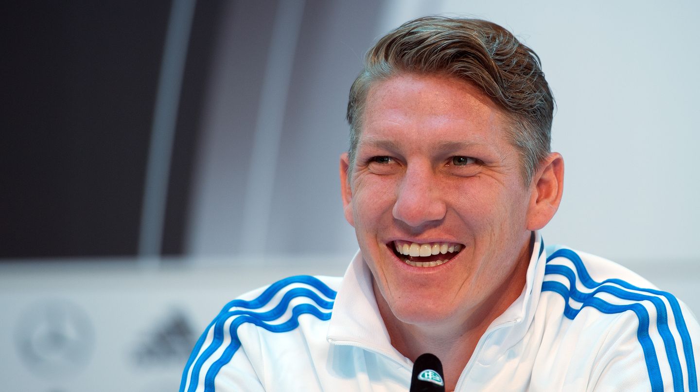 Bastian Schweinsteiger sitzt in weiß-blauer Trainingsjacke vor einem Mikrofon und grinst