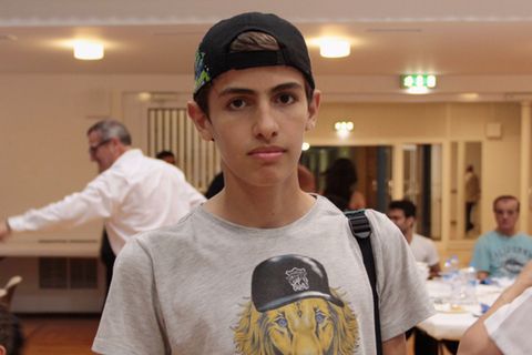 Bashir Salloum steht mit Kappe auf dem Kopf und einem Rucksackriemen an einer Schulter in einem Gemeinschaftsraum.