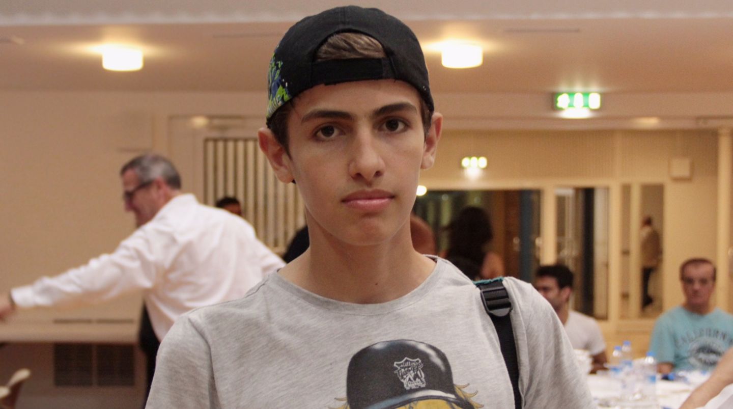 Bashir Salloum steht mit Kappe auf dem Kopf und einem Rucksackriemen an einer Schulter in einem Gemeinschaftsraum.