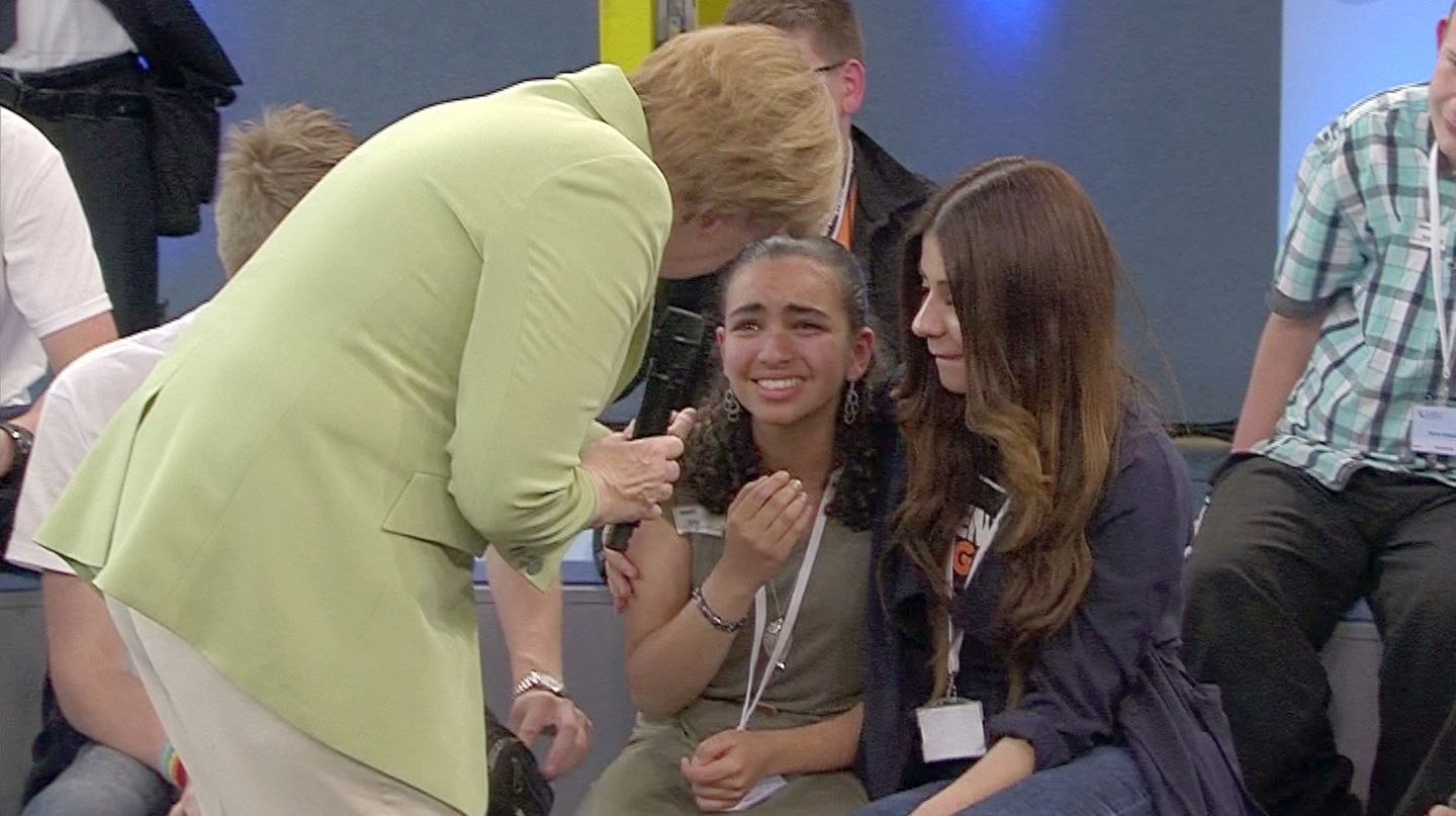 Angela Merkel im grünen Blazer beugt sich zu einem weinenden Mädchen herunter, das ein anderes Mädchen im Arm hält.