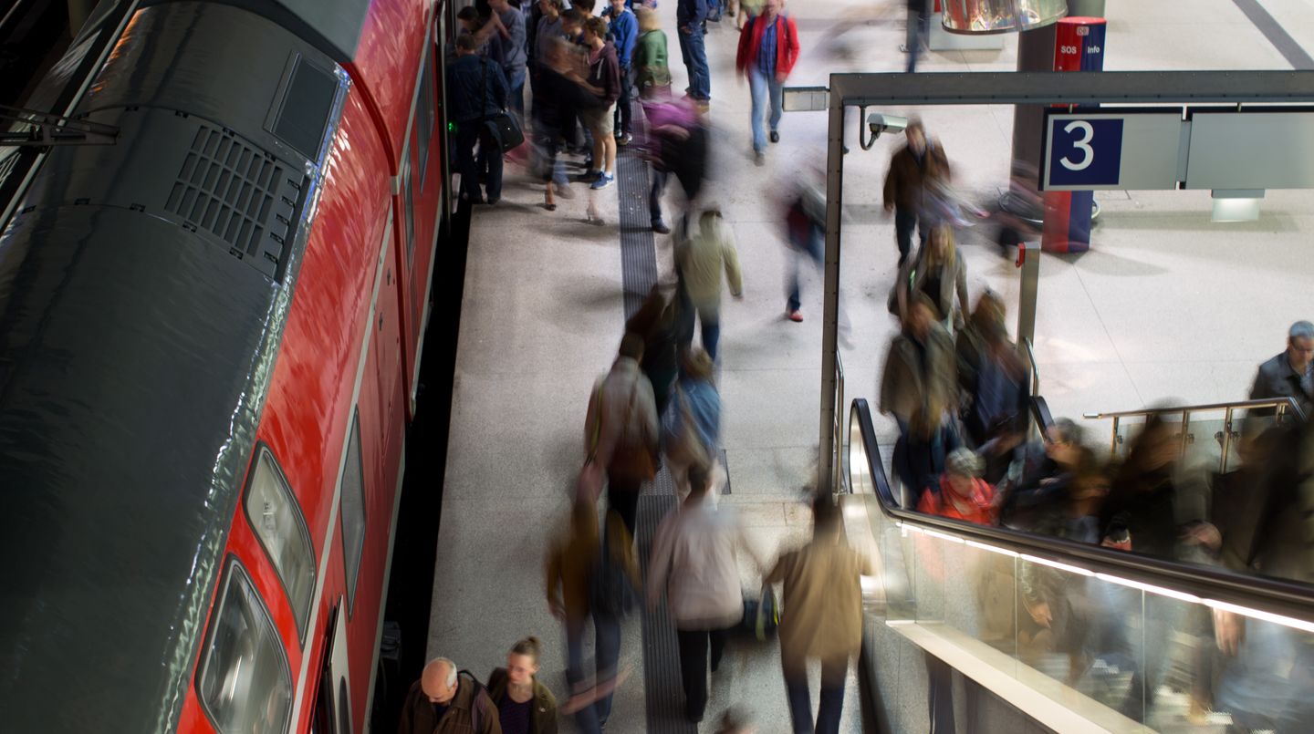An einem Bahngleis strömen Menschen - verzerrt unkenntlich gemacht - hin und her, auf die Rolltreppe, in einen Zug hinein.