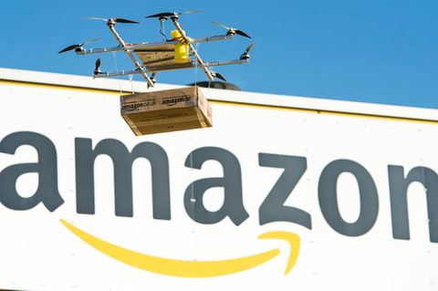 Amazon Prime Air: Eine Lieferdrohne mit einem Paket fliegt vor dem Amazon-Logo