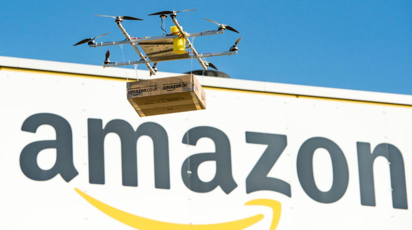Amazon Prime Air: Eine Lieferdrohne mit einem Paket fliegt vor dem Amazon-Logo