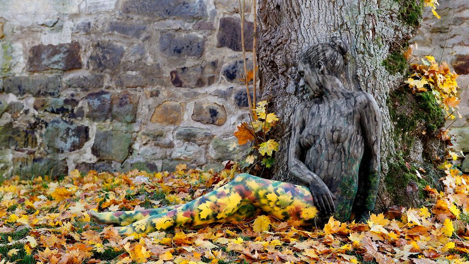 Frau sitzt mit Bodypainting kaum sichtbar im Laub, angelehnt an einen Baum