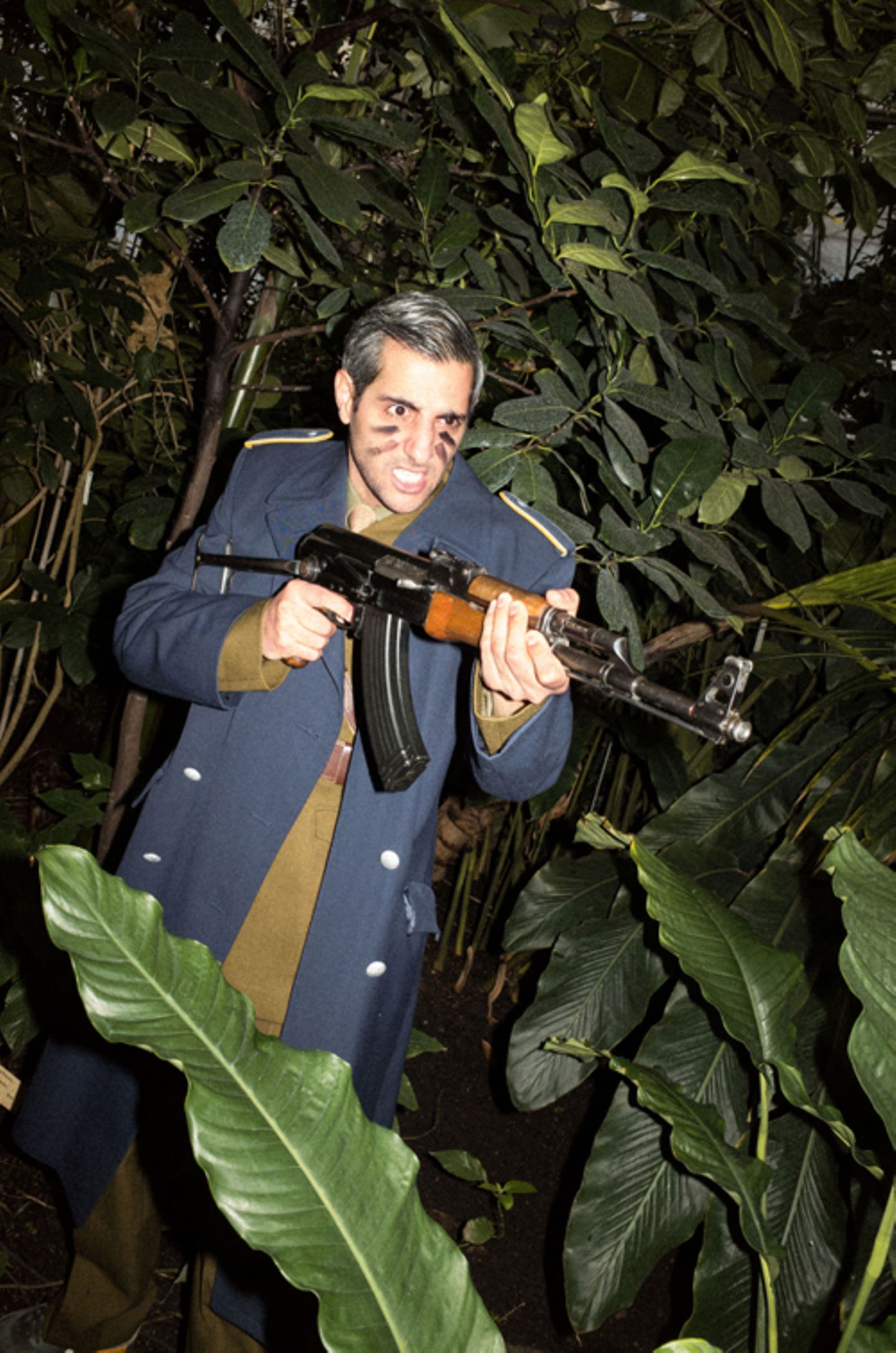 Michel Abdollahi steht in Uniform zwischen großen Grünpflanzen und hält ein Gewehr in der Hand.
