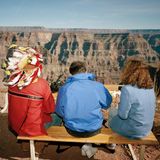 Auf einer Besucherbank am Grand Canyon in Arizona, USA, sitzten zwei Touristen neben einem Mann in partieller Indianermontur