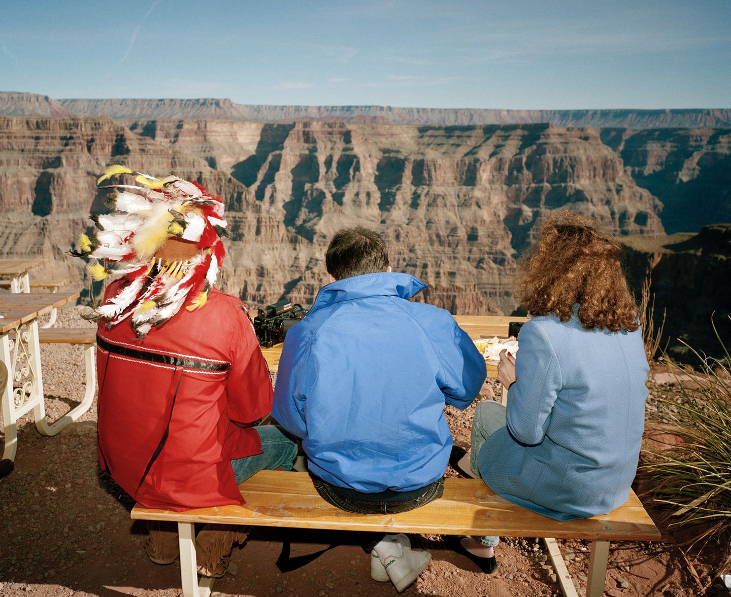 Auf einer Besucherbank am Grand Canyon in Arizona, USA, sitzten zwei Touristen neben einem Mann in partieller Indianermontur