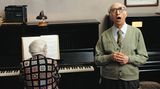 Eine alte Dame am Klavier, ein alter Herr singt. Über dem Klavier hängt eine Inschrift.