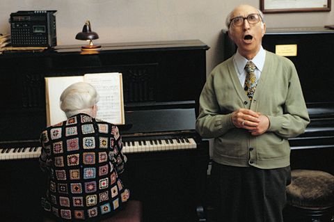 Eine alte Dame am Klavier, ein alter Herr singt. Über dem Klavier hängt eine Inschrift.