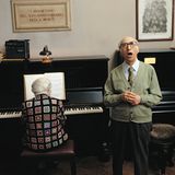 Eine alte Dame am Klavier, ein alter Herr singt. Über dem Klavier hängt eine Inschrift.