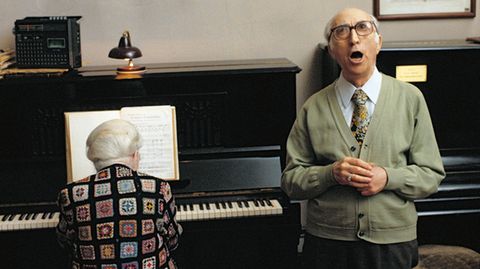 Eine alte Dame am Klavier, ein alter Herr singt. Über dem Klavier hängt eine Inschrift.