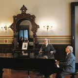 Ein älterer Herr am Flügel, ein weiterer und eine Dame singen dazu