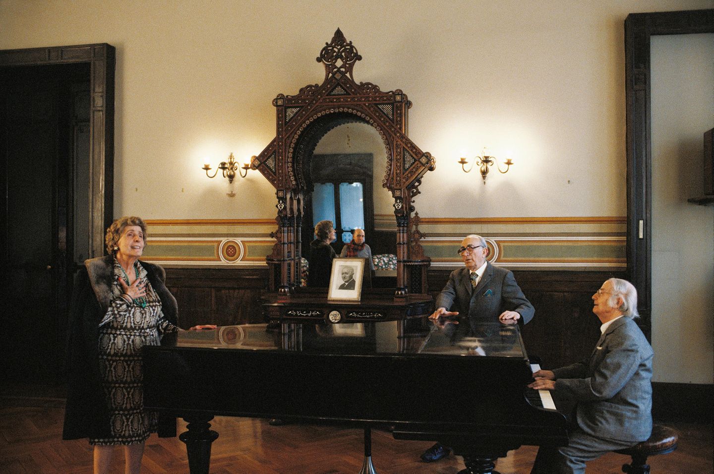 Ein älterer Herr am Flügel, ein weiterer und eine Dame singen dazu
