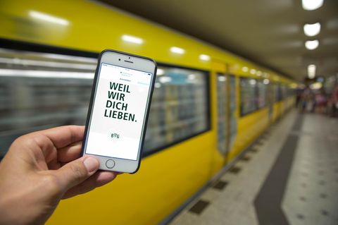 Ein Smartphone mit dem Schriftzug "Weil wir dich lieben" vor einer gelben Berliner U-Bahn