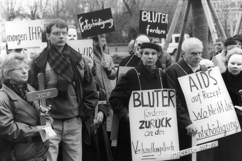 Zu sehen ist eine Demo mit mehreren Menschen, die Schilder emporstrecken, auf denen beispielsweise "Entschädigung" steht.