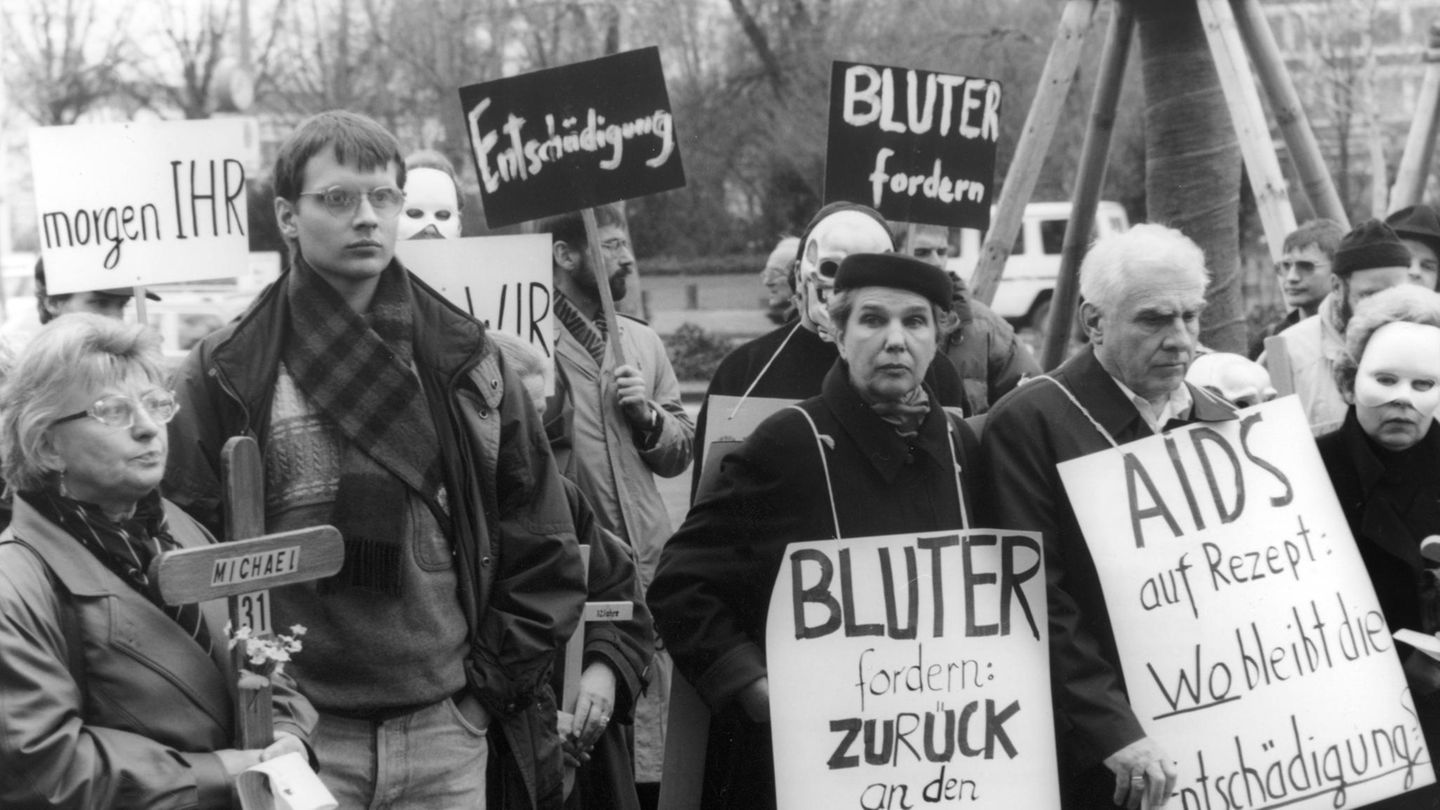 Zu sehen ist eine Demo mit mehreren Menschen, die Schilder emporstrecken, auf denen beispielsweise "Entschädigung" steht.