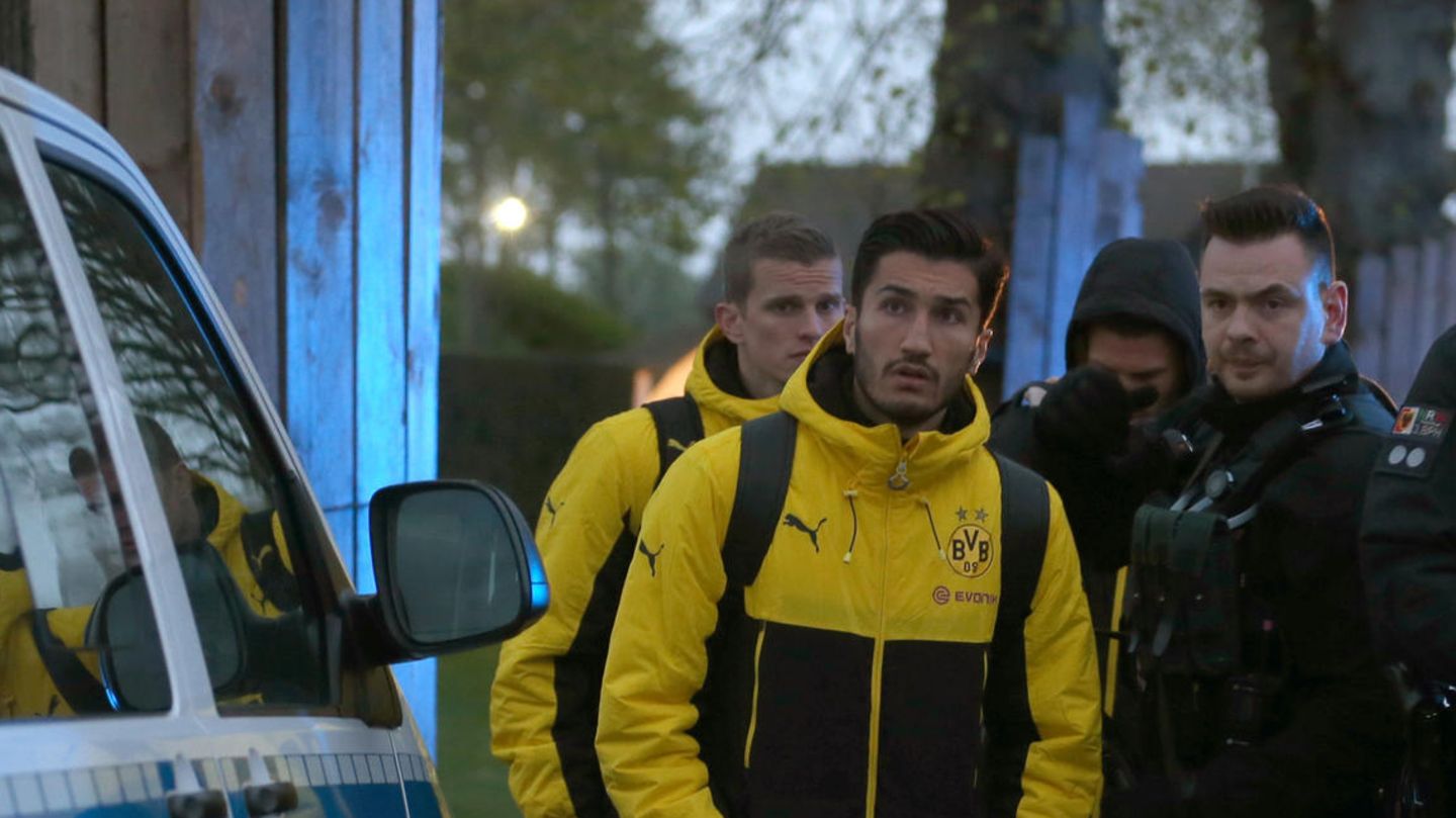 BVB-Spieler Nuri Sahin steht in Teamkleidung zwischen einem Poliziebus und bewaffneten Polizisten