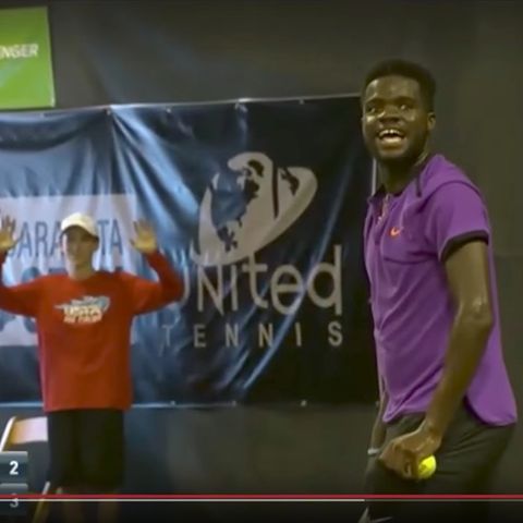 Tennisspieler Frances Tiafoe steht beim eigenen Aufschlag mit dem Ball in der Hand und muss lachen.