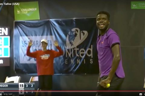 Tennisspieler Frances Tiafoe steht beim eigenen Aufschlag mit dem Ball in der Hand und muss lachen.