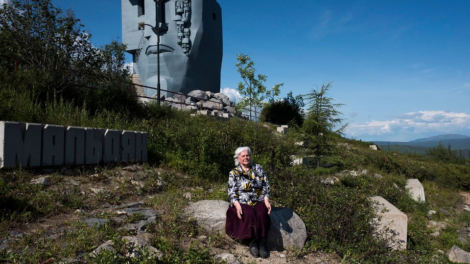 Die Gulag-Überlebende Stefanija vor der "Maske der Trauer" in Magadan, sitzend, vor einer grünen, abschüssigen Landschaft
