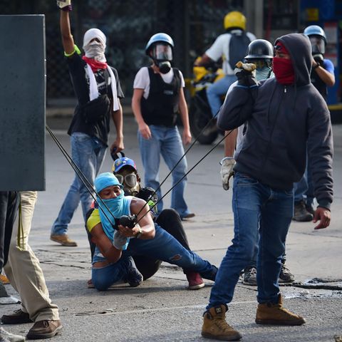 Drei vermummte junge Männer in Venezuela spannen eine Schleuder, um etwas auf Polizisten zu schießen