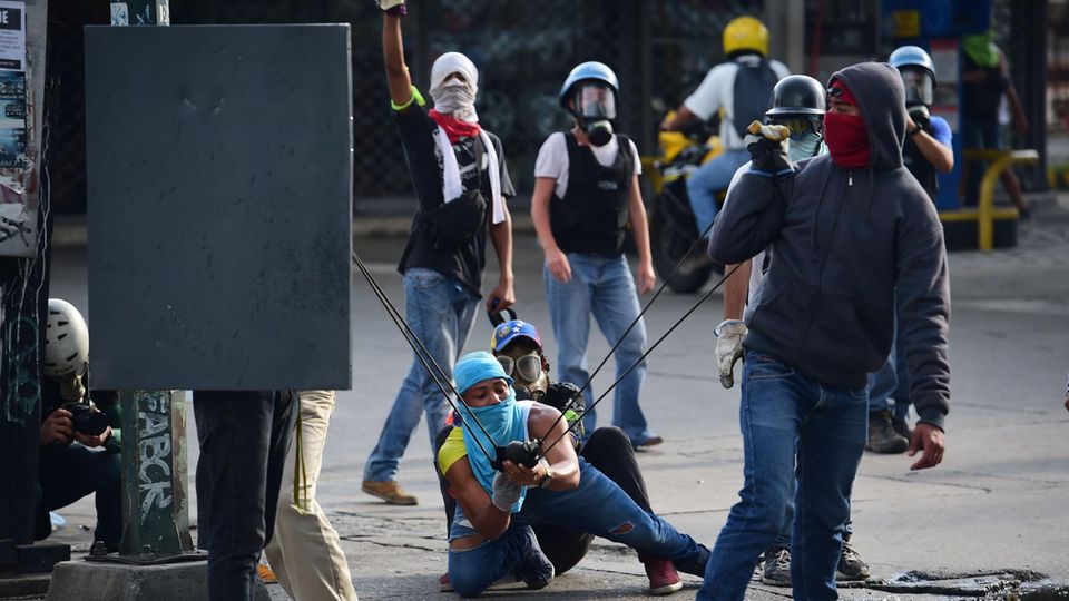 Drei vermummte junge Männer in Venezuela spannen eine Schleuder, um etwas auf Polizisten zu schießen