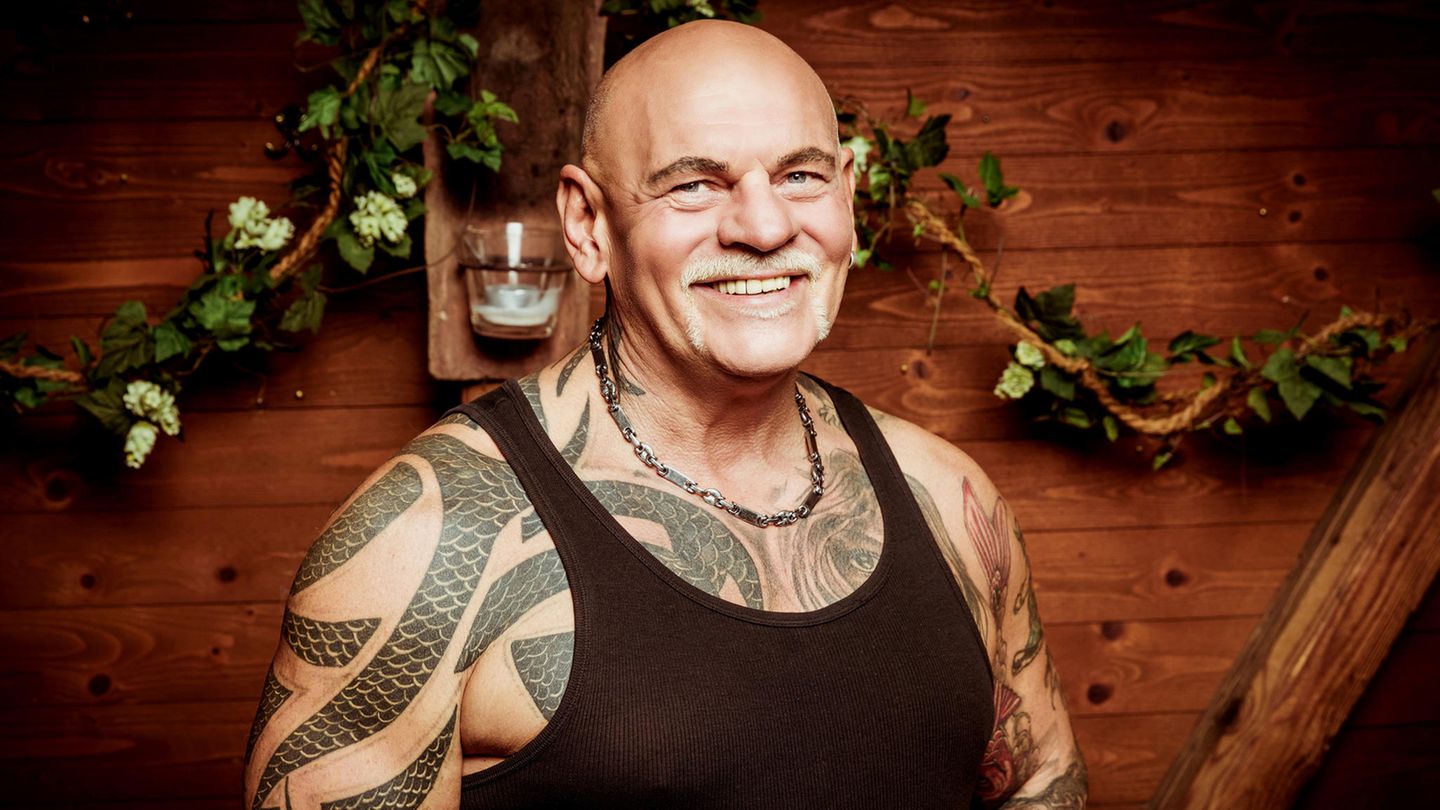 Alfred, ein kräftiger Mann mit Glatze und Tattoos unter dem schwarzen Unterhemd, lächelt vor einer Holzwand in die Kamera