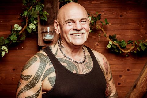 Alfred, ein kräftiger Mann mit Glatze und Tattoos unter dem schwarzen Unterhemd, lächelt vor einer Holzwand in die Kamera