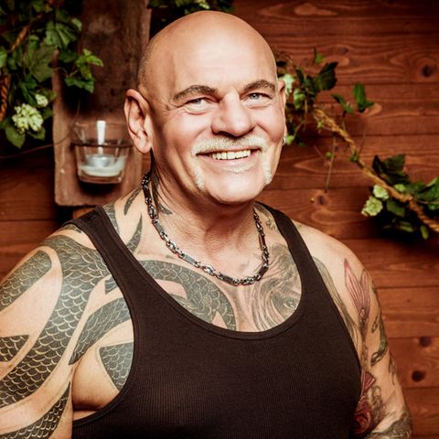 Alfred, ein kräftiger Mann mit Glatze und Tattoos unter dem schwarzen Unterhemd, lächelt vor einer Holzwand in die Kamera