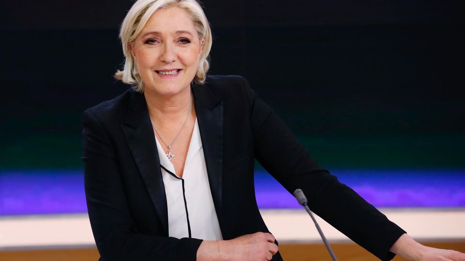 Marine Le Pen sitzt in einem Fernsehstudio vor einer blauschwarzen Wand und lächel