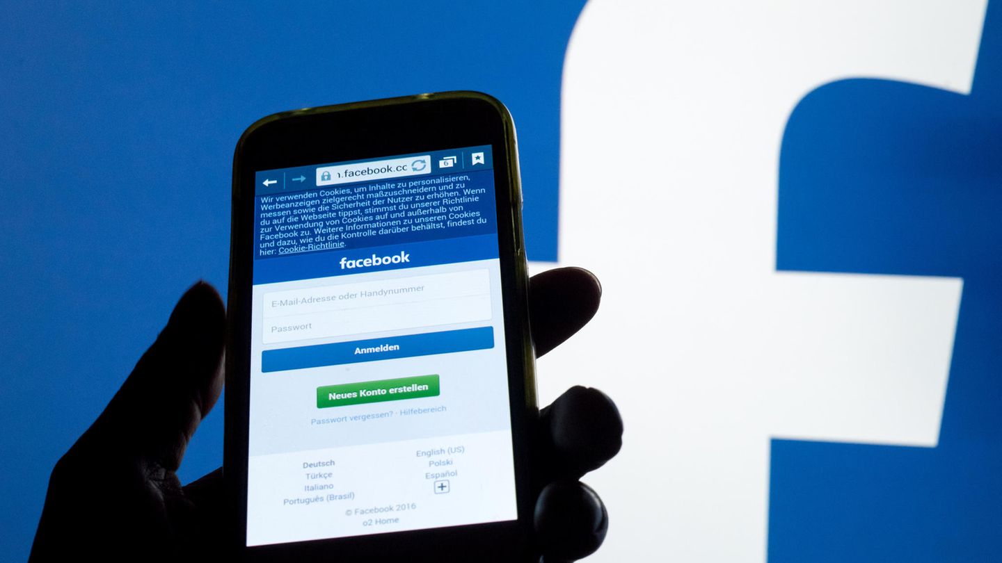 Ein Smartphone Display mit der Login-Seite von Facebook leuchtet vor einer Wand mit einem großen Facebook-Logo