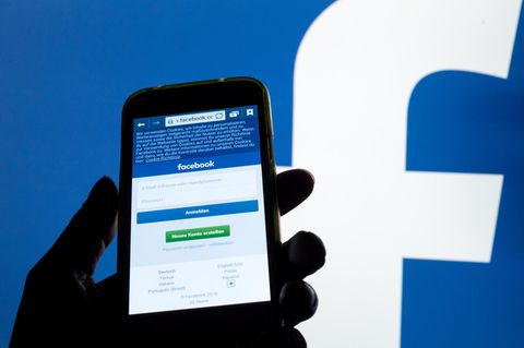 Ein Smartphone Display mit der Login-Seite von Facebook leuchtet vor einer Wand mit einem großen Facebook-Logo