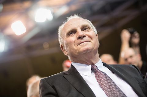 Uli Hoeneß sitzt im Anzug auf der Jahreshauptversammlung des FC Bayern München und lächelt etwas verkniffen