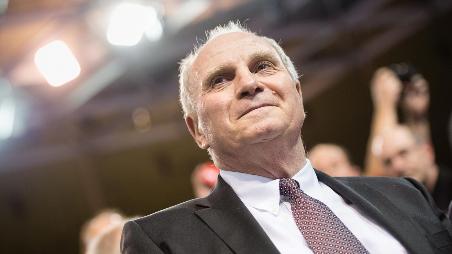Uli Hoeneß sitzt im Anzug auf der Jahreshauptversammlung des FC Bayern München und lächelt etwas verkniffen