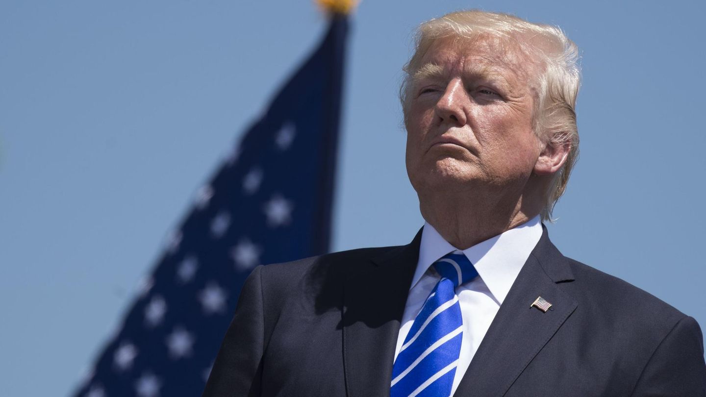 US-Präsident Donald Trump steht unter blauem Himmel im Freien, Sonne scheint ihm ins Gesicht. Hinter ihm weht eine US-Flagge