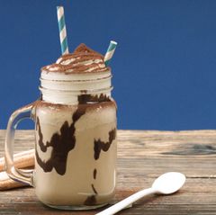 Aus einem Trinkglas mit Sahnehaube ragen zwei blau-weiß geringelte Strohhalme heraus. Im Glas eine kakaofarbene Flüssigkeit.