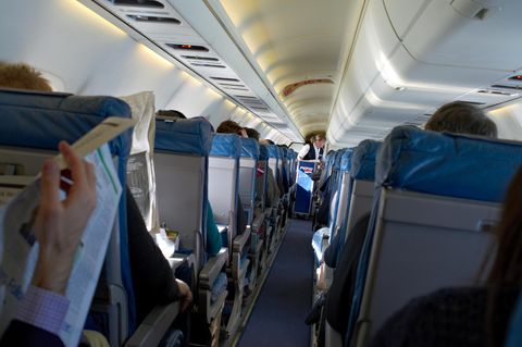 Fluggäste sitzen an Bord eines Flugzeuges, während eine Flugbegleiterin im Gang Getränke verteilt