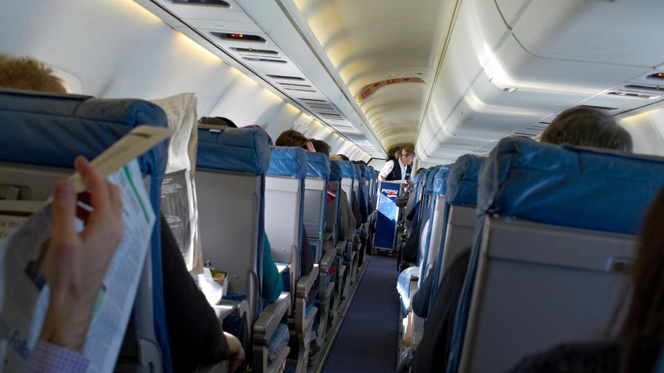 Fluggäste sitzen an Bord eines Flugzeuges, während eine Flugbegleiterin im Gang Getränke verteilt