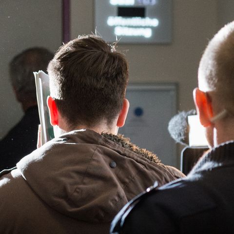 Ein Mann im Parka geht links und verbirgt sein Gesicht mit einem Ordner. Rechts hinter ihm geht ein Justizbeamter.