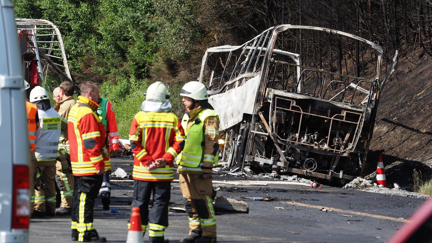 Busunglück auf der A9 in Bayern: Der ausbrannte Bus liegt am Straßenrand, davor stehen Einsatzkräfte der Feuerwehr