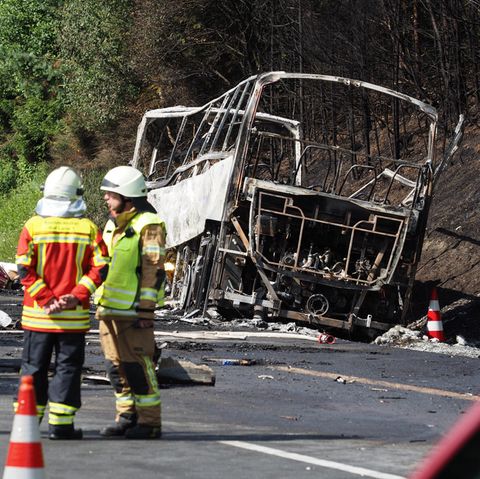 Busunglück auf der A9 in Bayern: Der ausbrannte Bus liegt am Straßenrand, davor stehen Einsatzkräfte der Feuerwehr