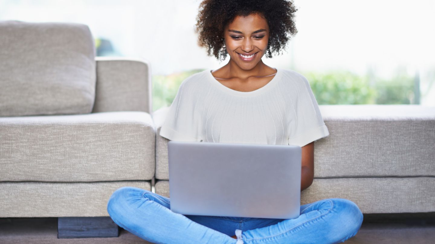 Eine Frau sitzt zuhause mit dem Laptop auf dem Boden und lächelt