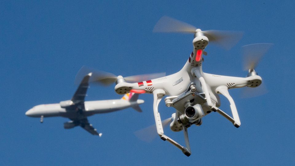Eine private Drohne fliegt in knapp 10 Metern Flughöhe über dem Boden, während in weiter Entfernung ein Flugzeug zu sehen ist.